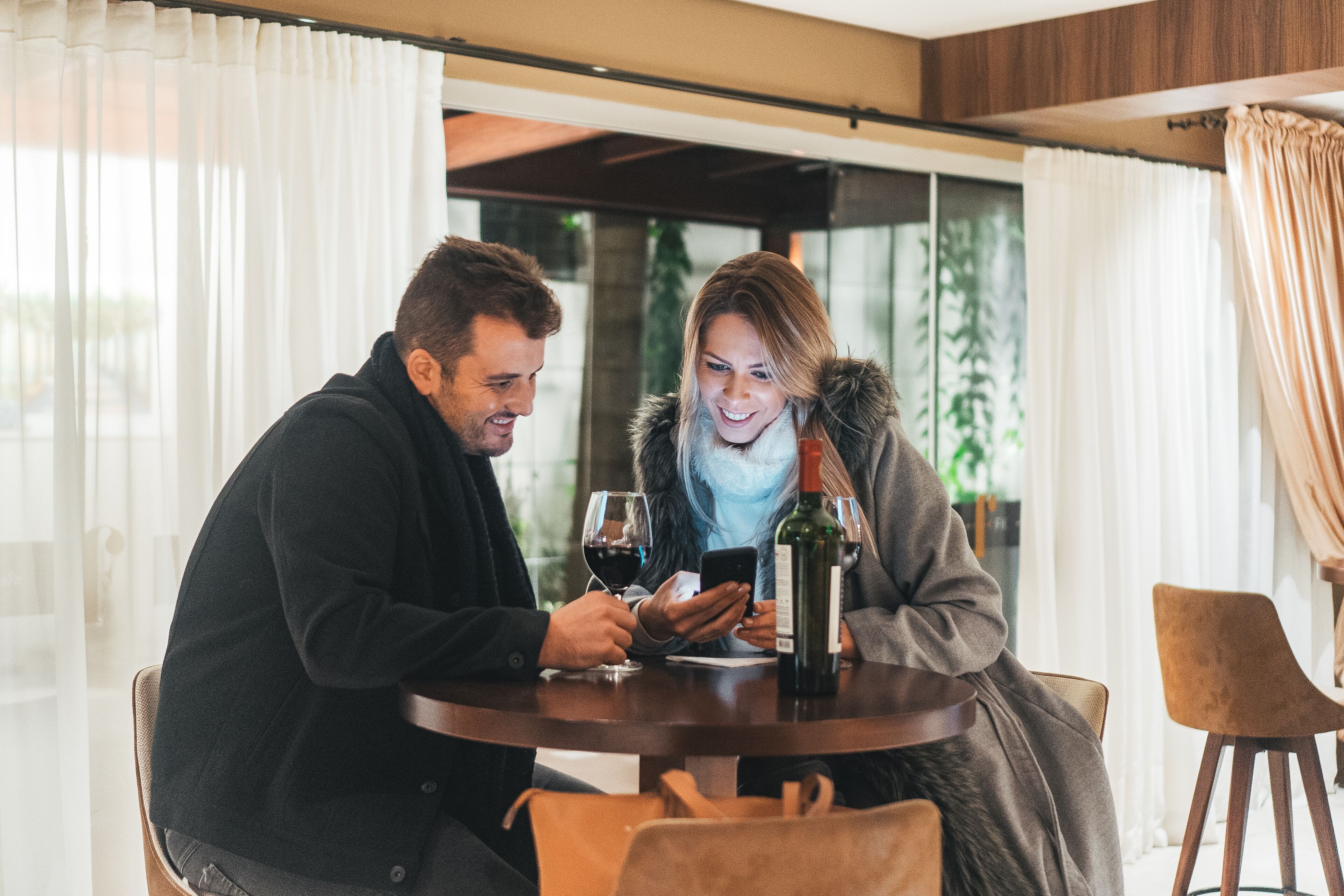 Un couple est assis à une table ronde, devant une bouteille de vin et des verres, dans une pièce dotée de grandes fenêtres et de rideaux.