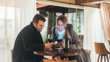 Un couple est assis à une table ronde, devant une bouteille de vin et des verres, dans une pièce dotée de grandes fenêtres et de rideaux.