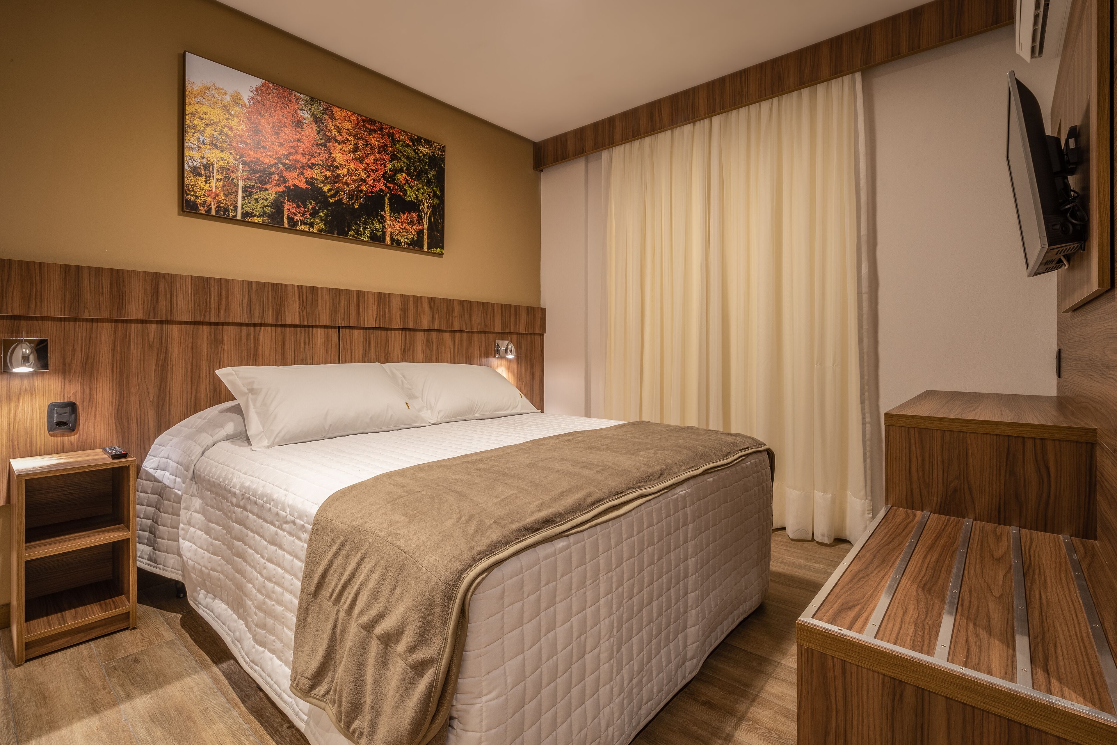 A hotel room with a large bed, wooden headboard, a bedside table with a lamp, a TV mounted on the wall, and a window with curtains.