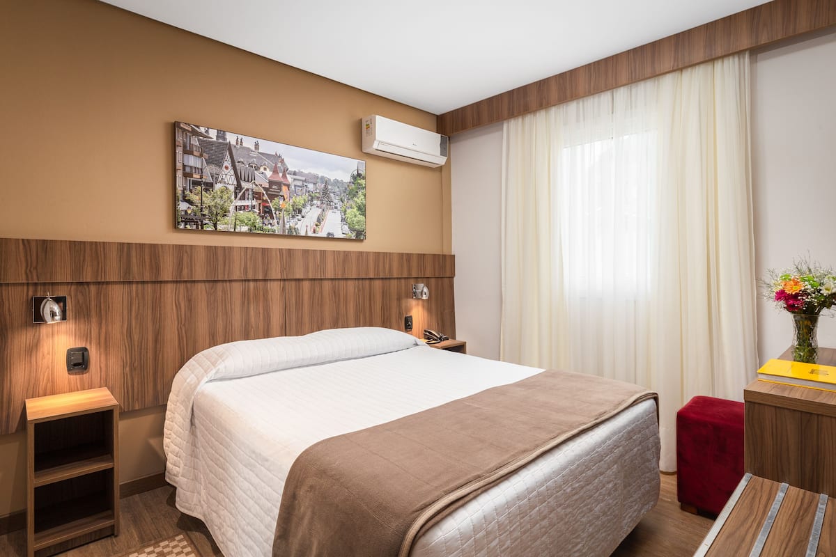 A hotel room with a large bed, a wooden headboard, a nightstand with a lamp, a small table with a vase of flowers, and a painting on the wall.