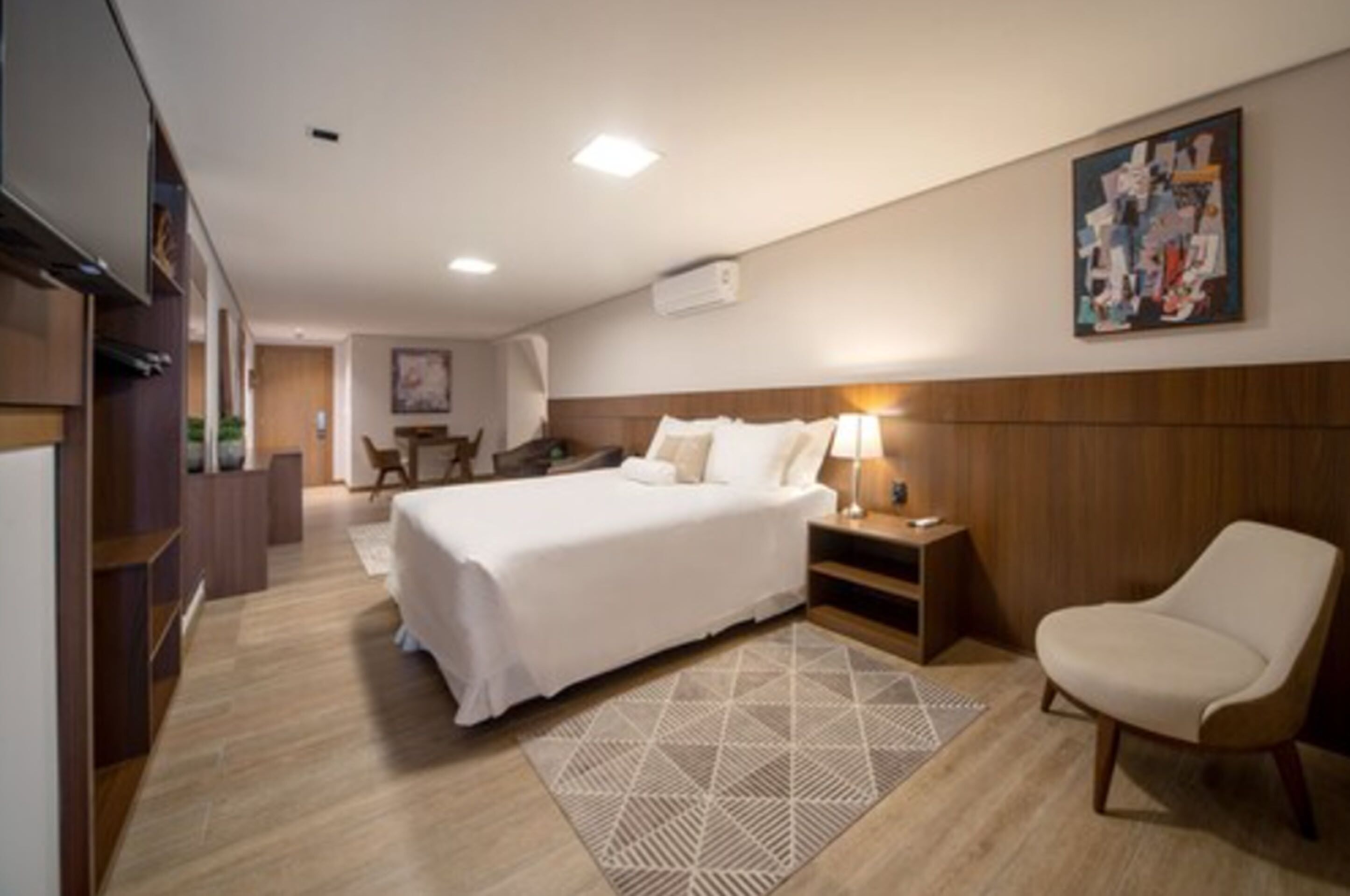 Una habitación de hotel con una cama grande, una mesita de noche con una lámpara, una silla, un cabecero de madera y un cuadro en la pared.