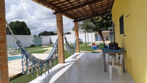 A covered patio area with a hammock, a table with chairs, and a pool in the background. - Chacara So Alegria em Parnamirim (Parnamirim)