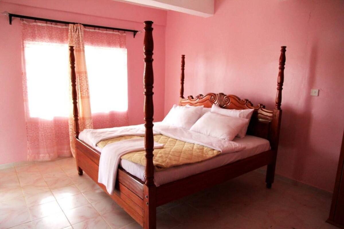 Uma cama de dossel em um quarto com paredes cor-de-rosa e uma janela com cortinas leves.