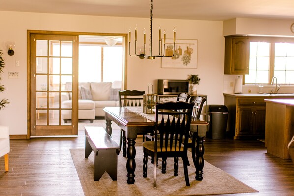 A dining area with a wooden table, chairs, and a chandelier. There is a kitchen in the background with wooden cabinets and a window.