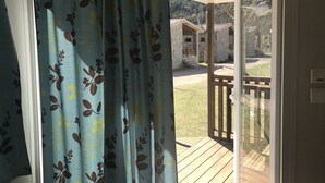 A view through a curtain with a floral pattern, revealing a wooden deck, stone buildings, and a grassy area. - Camping Pirinenc (Campdevanol)