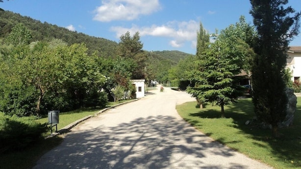 A rural road leading to a white building with a small entrance, surrounded by lush greenery and trees. - Camping Pirinenc (Campdevanol)