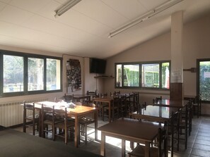 A dining area with multiple wooden tables and chairs, large windows with views of greenery, a bulletin board with photos, and a television mounted on the wall. - Camping Pirinenc (Campdevanol)