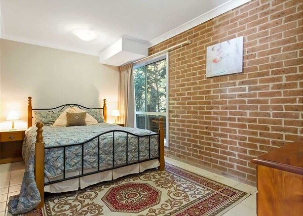 A bedroom with a bed, a nightstand, a wooden dresser, a window, and a brick wall.