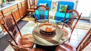 Un coin repas avec une table en verre entourée de chaises en bois, un canapé bleu agrémenté de coussins aux motifs tropicaux, et une cuisine en arrière-plan.