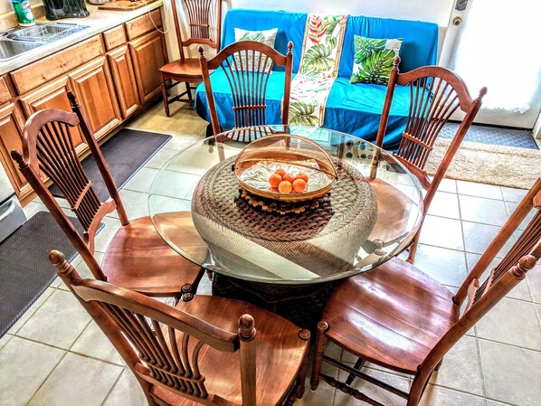 A dining area with a glass table surrounded by wooden chairs, a blue sofa with tropical pillows, and a kitchen in the background.