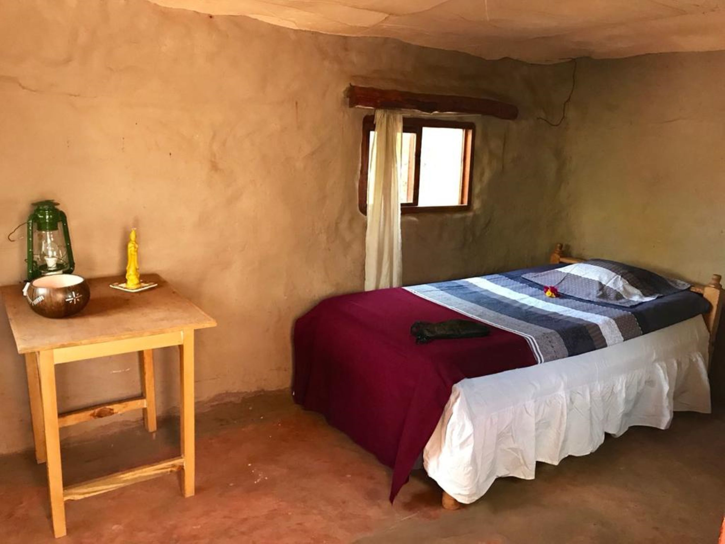 A room with a single bed, a wooden table, a green lantern, and a small statue.