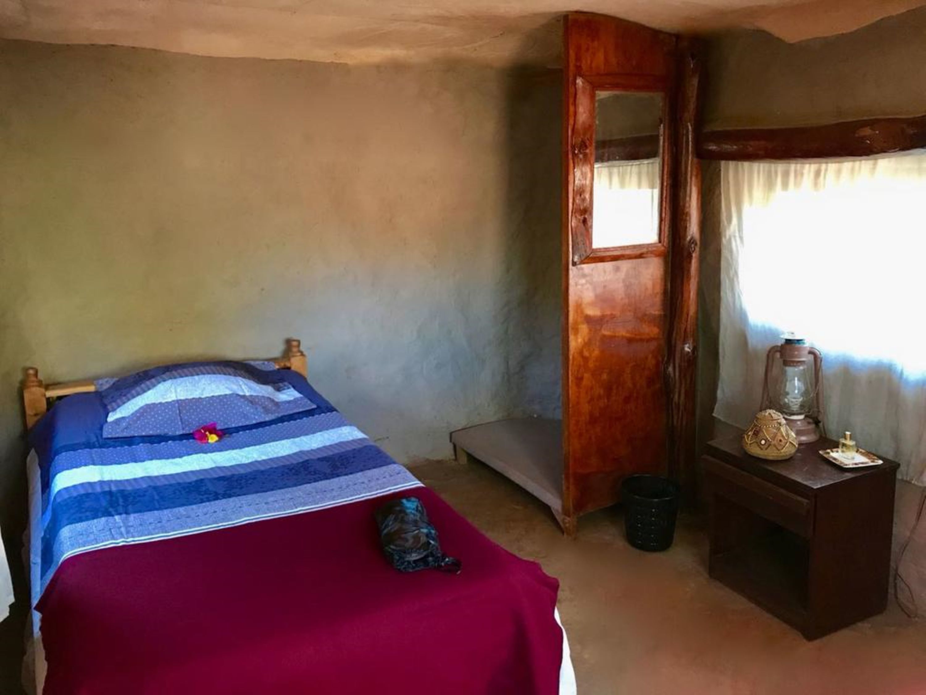 Ein Zimmer mit einem Bett, einer Holztür, einem kleinen Tisch mit einer Laterne und einem Fenster mit Vorhängen.