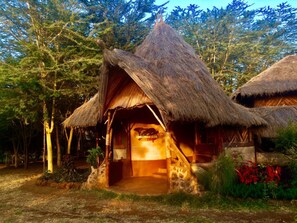 Uma cabana com telhado de palha, porta de madeira e uma placa com a inscrição 'AMBOSEI ECO'.