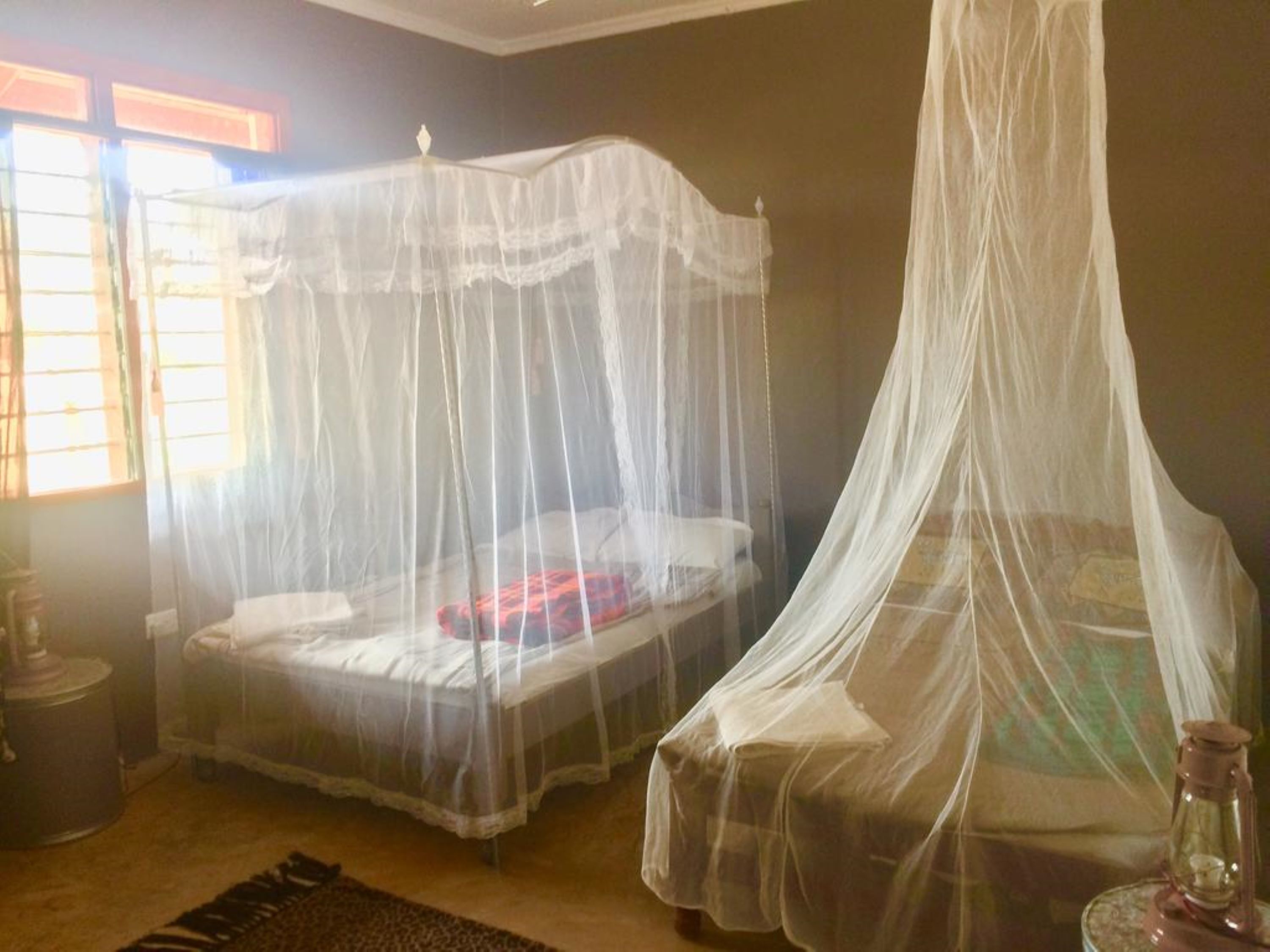 A room with two beds covered by white mosquito nets, a wooden floor, and a small table with a lantern.