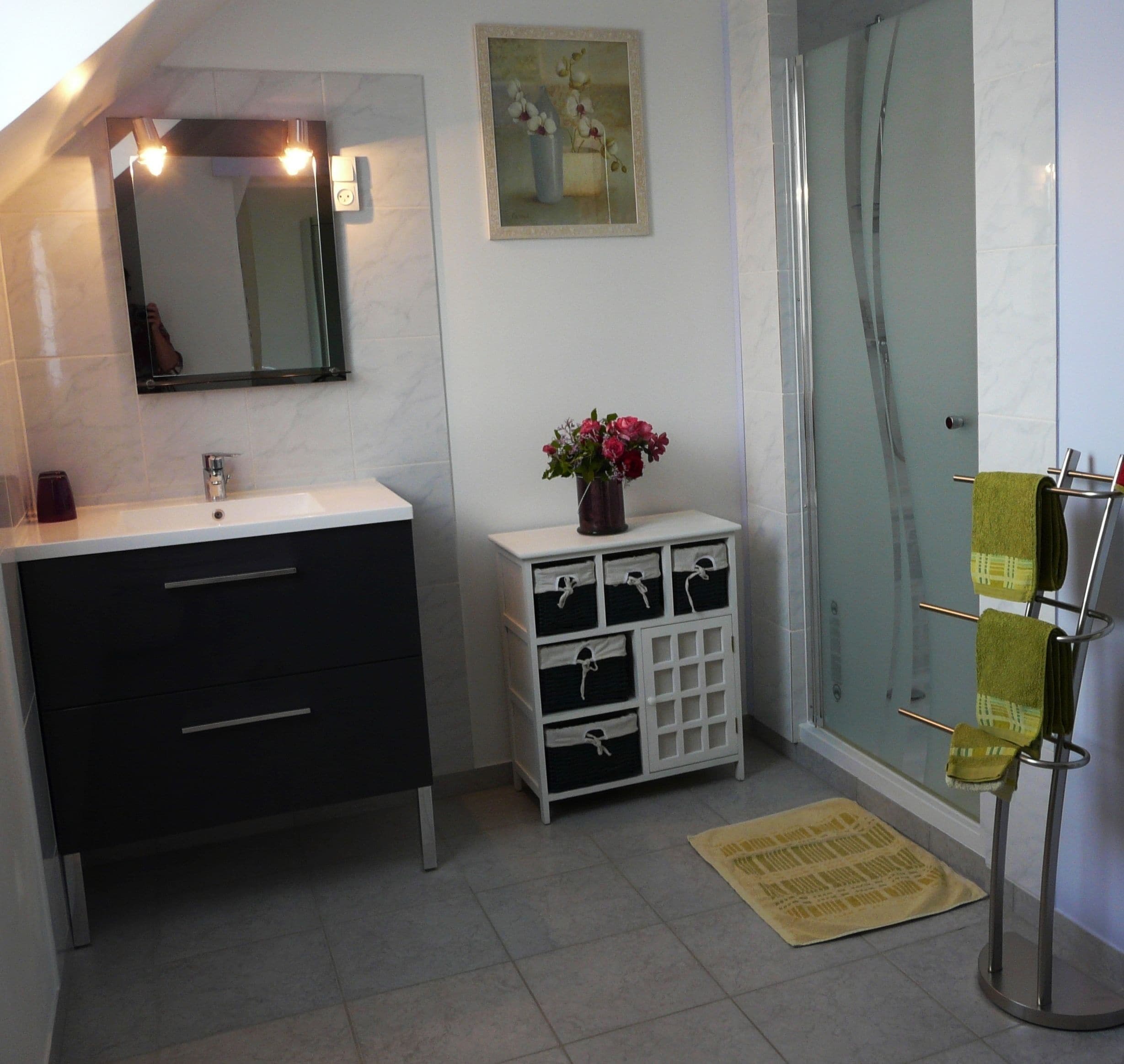 A bathroom with a white sink, dark cabinets, a mirror, a vase with flowers, a glass shower enclosure, and a towel rack.