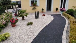 A building with a gravel pathway leading to a door, surrounded by potted plants and a small garden area. - Chambres d'hôtes le Pot au Lait (Villaines-la-Juhel)