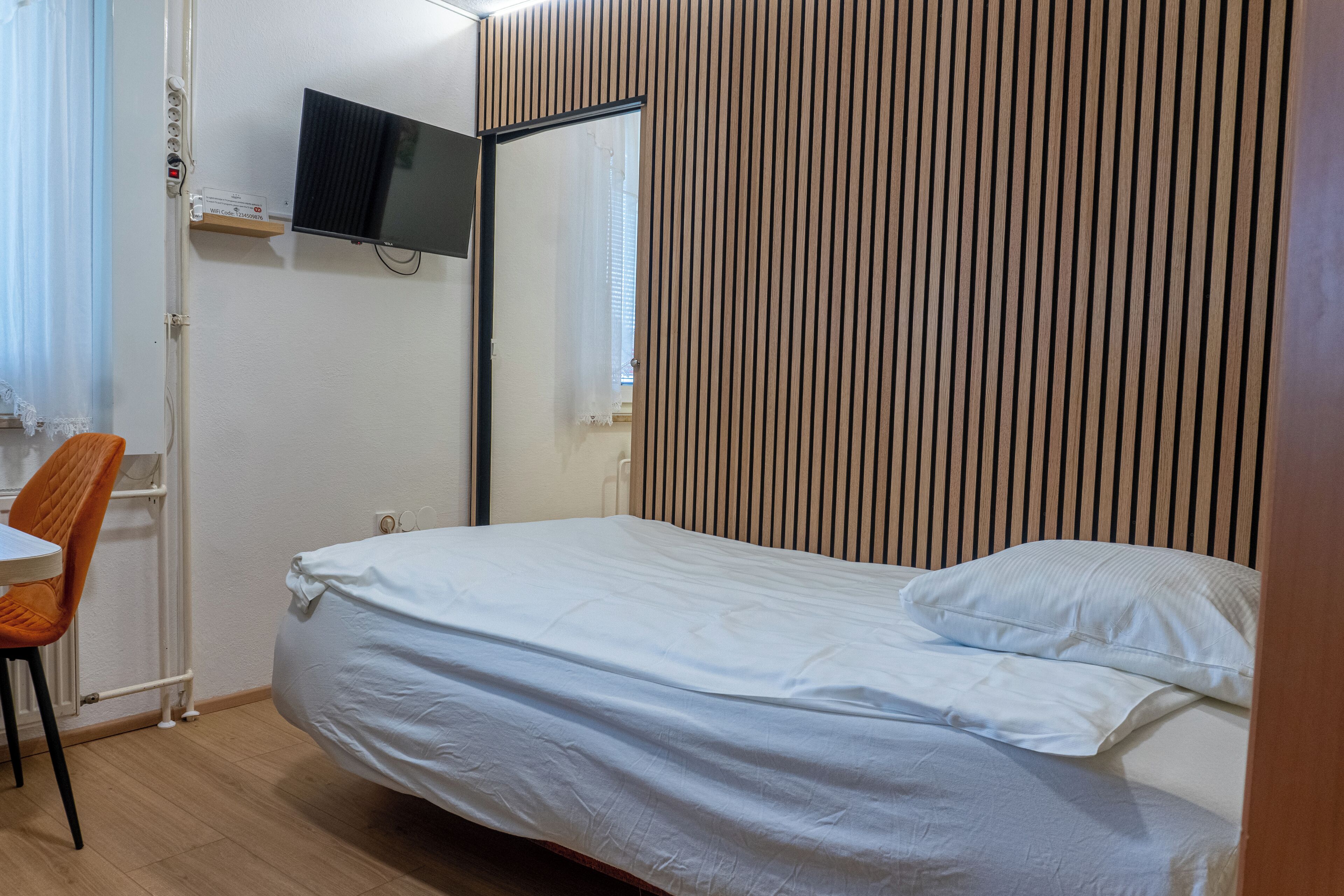 Een slaapkamer met een bed, een stoel, een aan de muur bevestigde televisie en een raam met gordijnen.