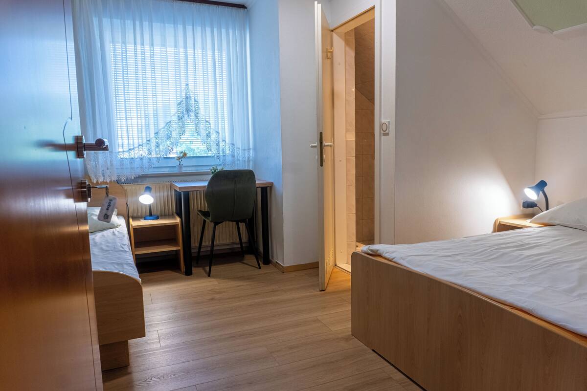 Ein Schlafzimmer mit Bett, Schreibtisch mit Stuhl, Fenster mit Vorhängen und einer Tür.