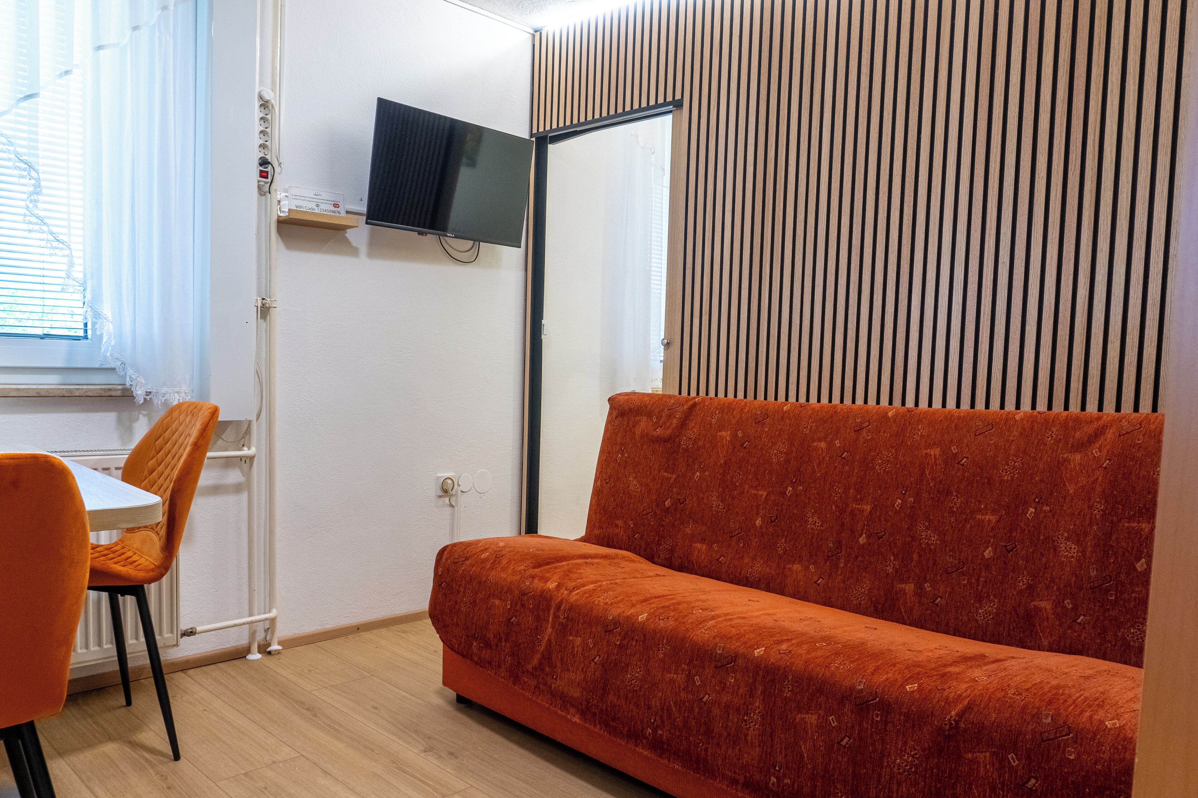 A room with a sofa, a TV mounted on the wall, a window with blinds, and a table with chairs.