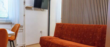 Una habitación con sofá, televisor en la pared, ventana con persianas y una mesa con sillas.