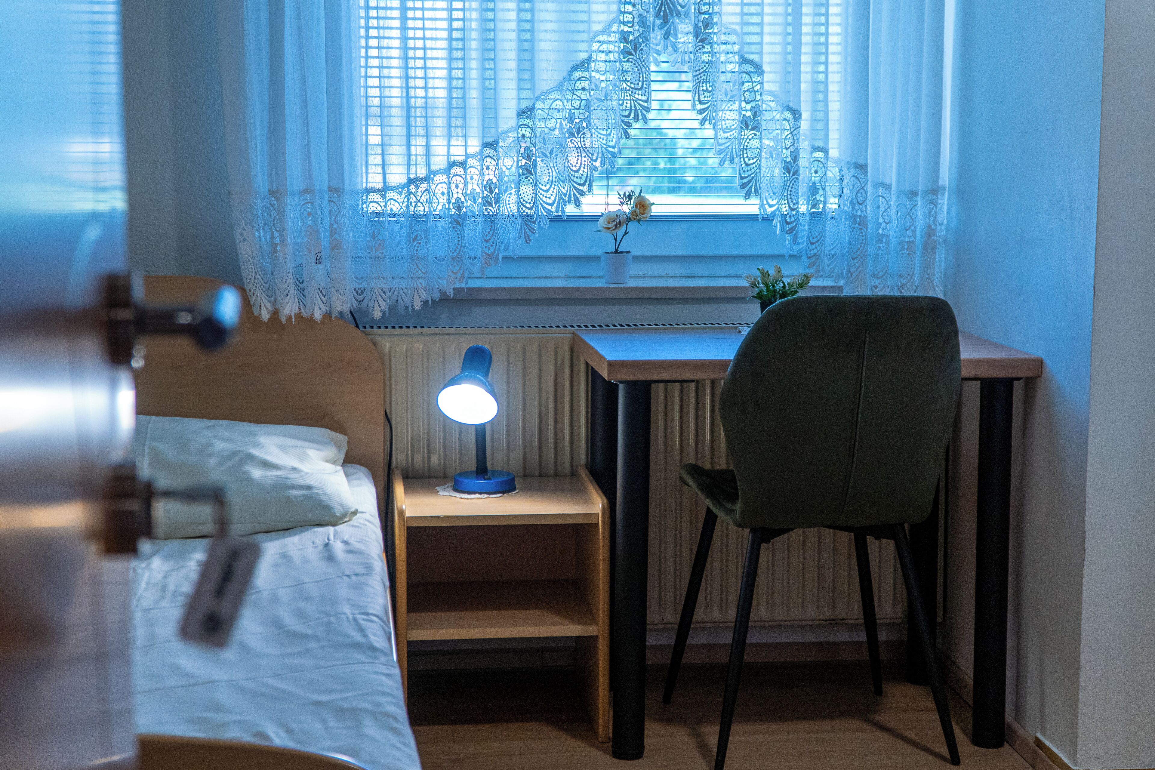 Ein Schlafzimmer mit Bett, Schreibtisch mit Stuhl, Nachttisch mit Lampe und einem Fenster mit Spitzenvorhängen.