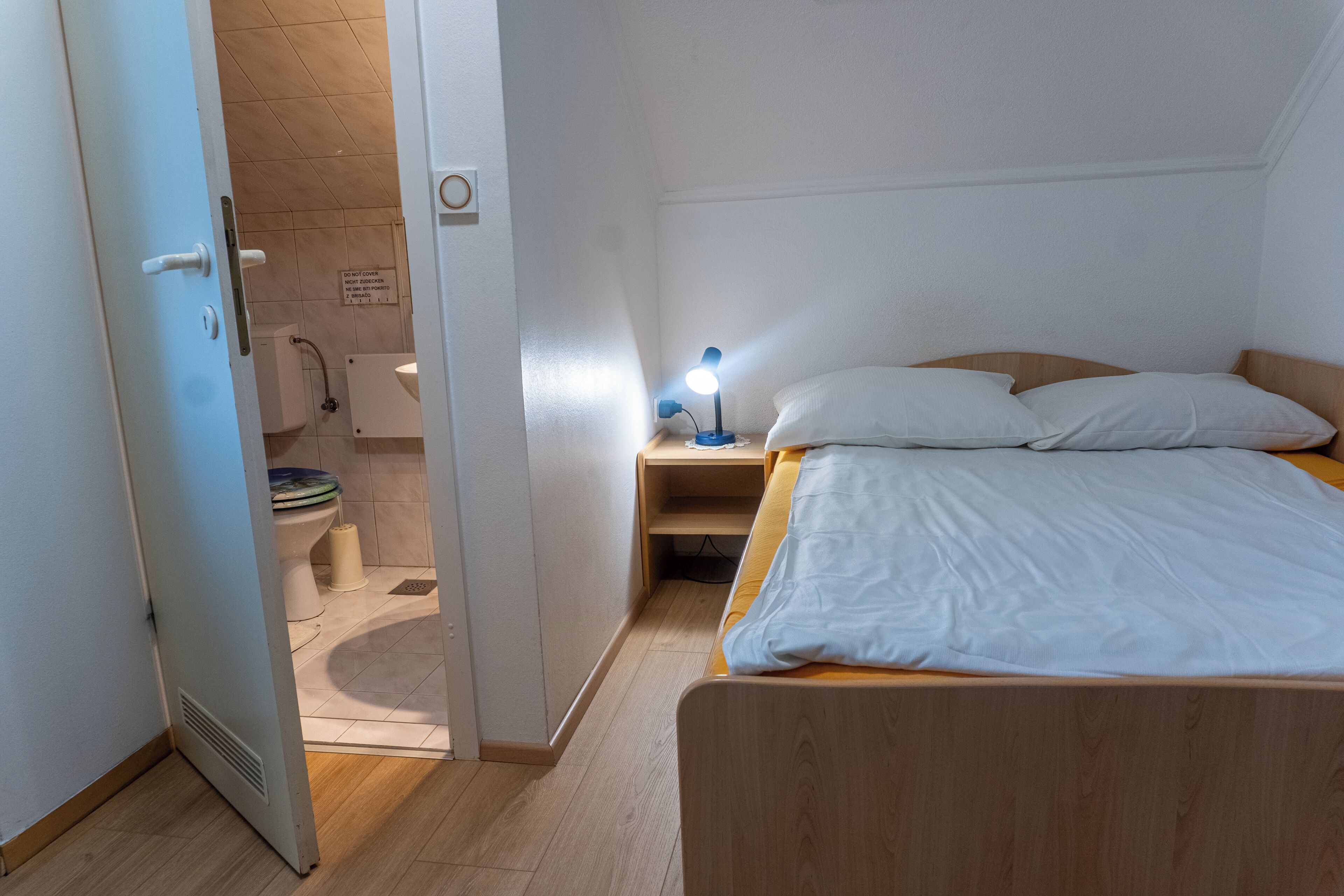 Een slaapkamer met een houten bed, een nachtkastje met een lamp, een witte deur en uitzicht op een badkamer met toilet en douche.