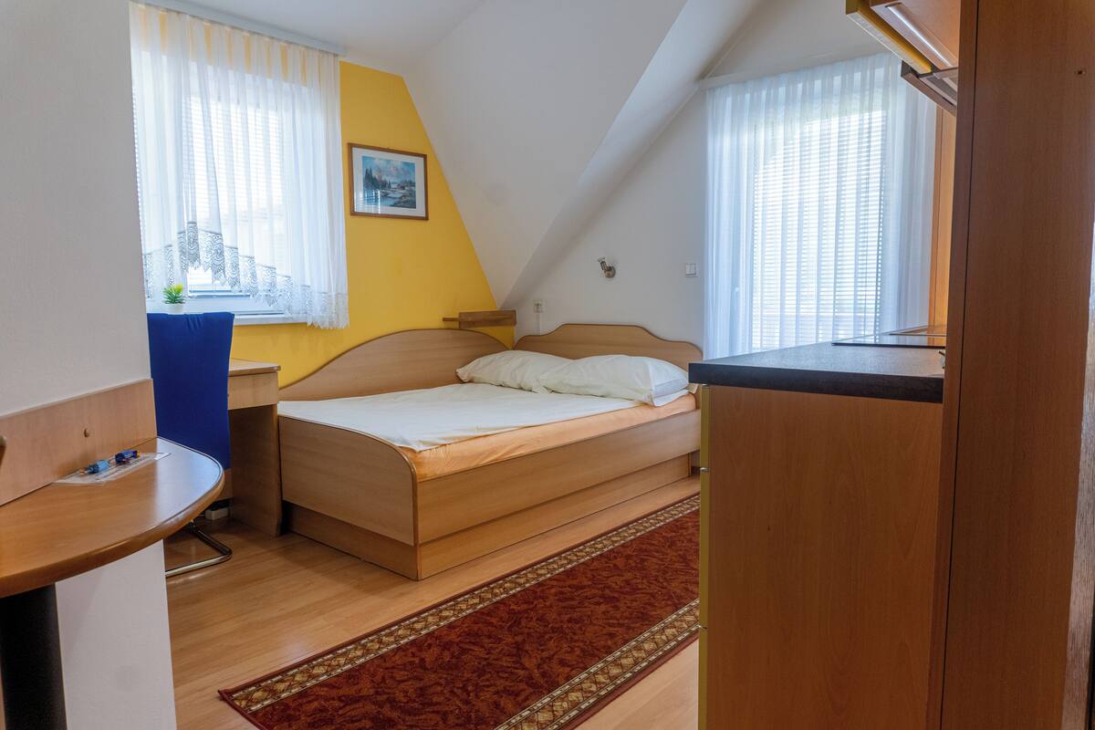 Ein Einzelbett mit Holz Kopfteil, ein kleiner Schreibtisch, ein Stuhl und ein Fenster mit durchsichtigen Vorhängen.