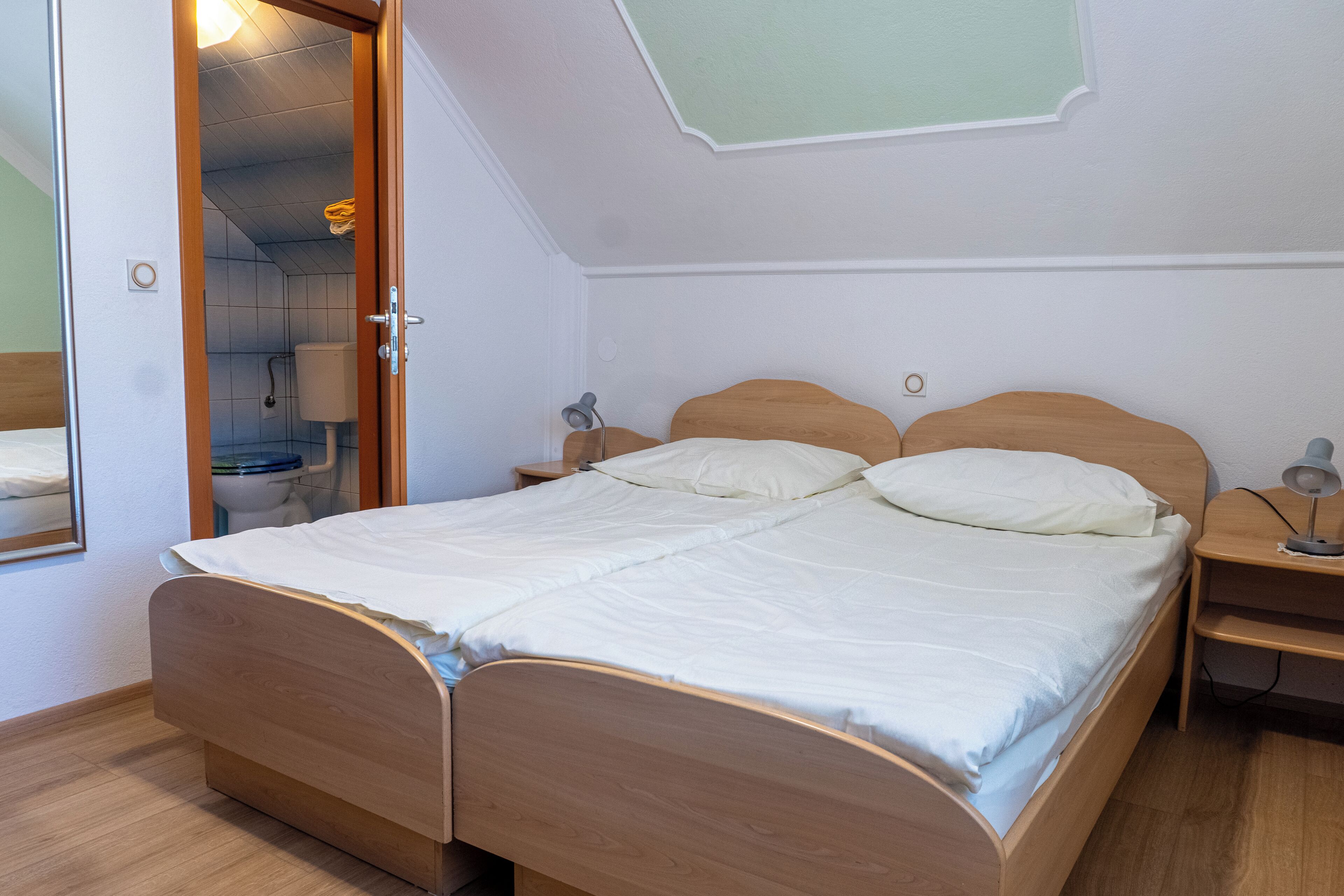 A bedroom with a wooden bed, a nightstand, a bedside lamp, and a bathroom visible through an open door.