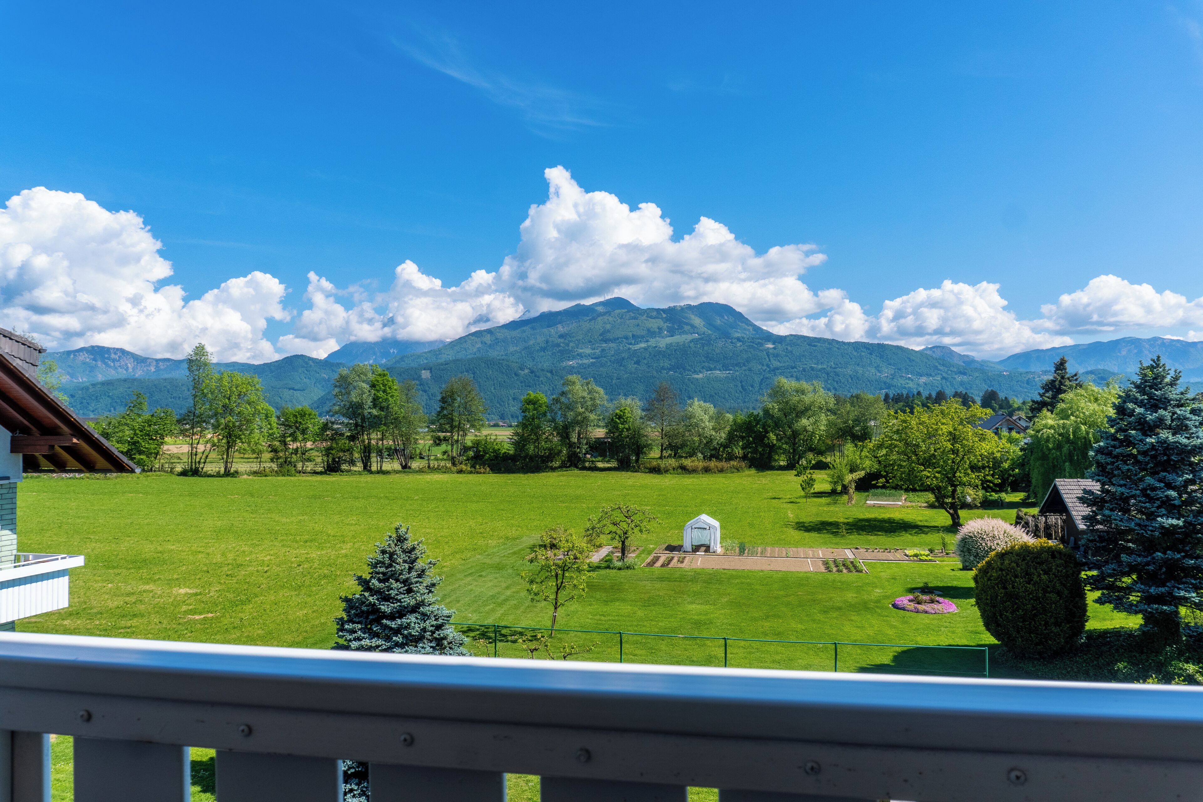 Uitzicht vanaf een balkon over een groen veld met een klein wit gebouw, bomen en bergen op de achtergrond.