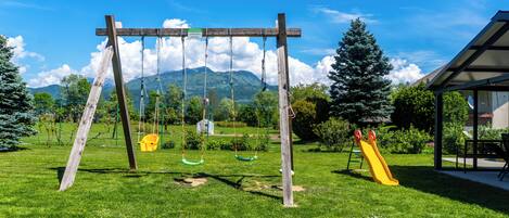 A playground with swings and a slide, surrounded by green grass and trees, with mountains in the background.