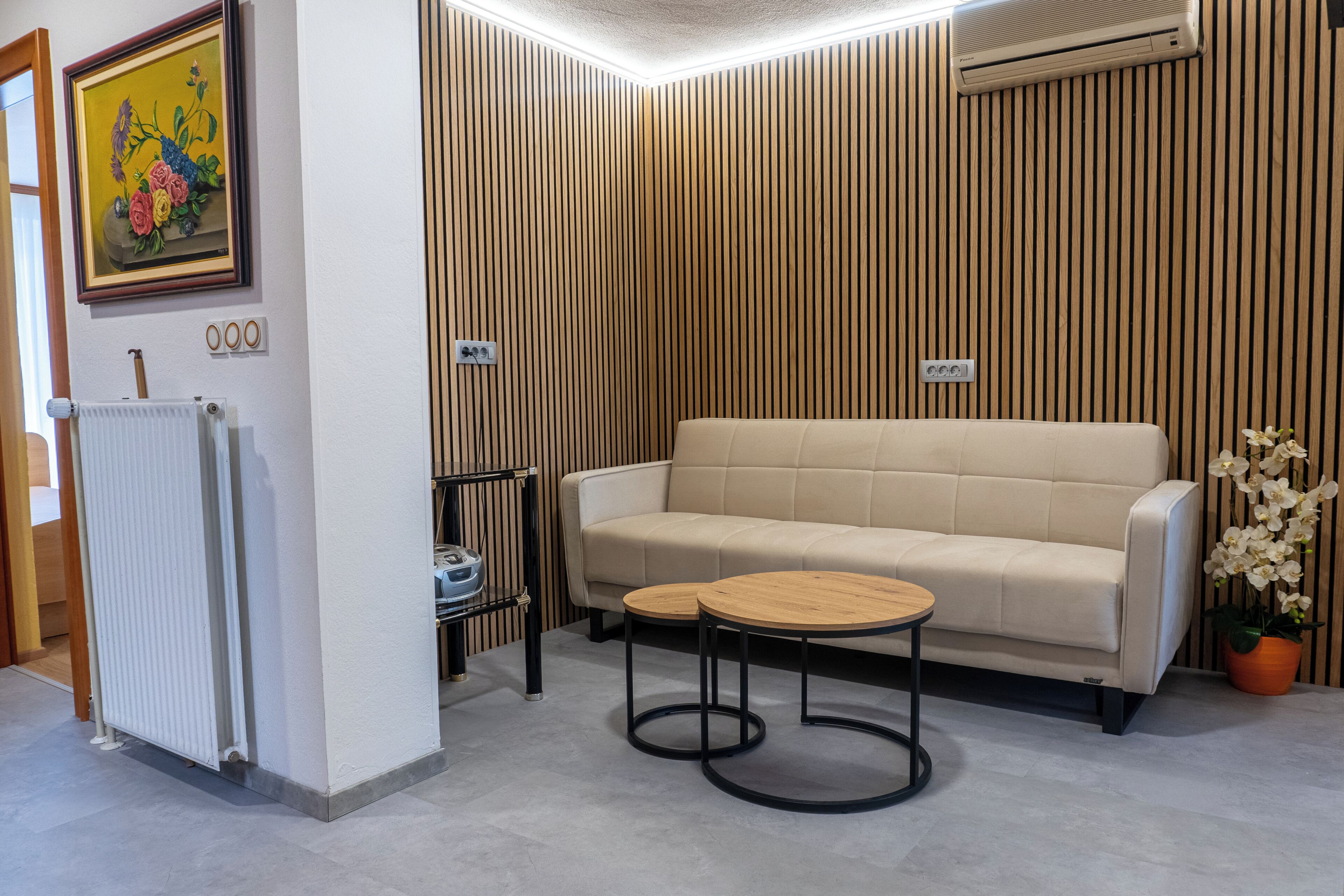 Una stanza con un divano color beige, due tavolini in legno, un termosifone, un quadro con fiori e un'orchidea in vaso.