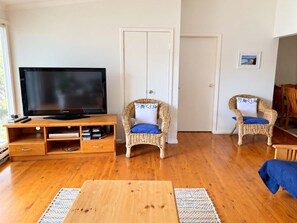 A living room with a flat-screen TV on a wooden stand, two wicker chairs with cushions, a wooden coffee table, and a door in the background.