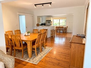 A spacious dining area with a wooden table and chairs, a kitchen in the background, and a view of the outdoors through large windows.