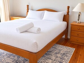 A neatly made bed with white linens and a rolled towel, a wooden nightstand with a lamp, and a patterned rug on a wooden floor.