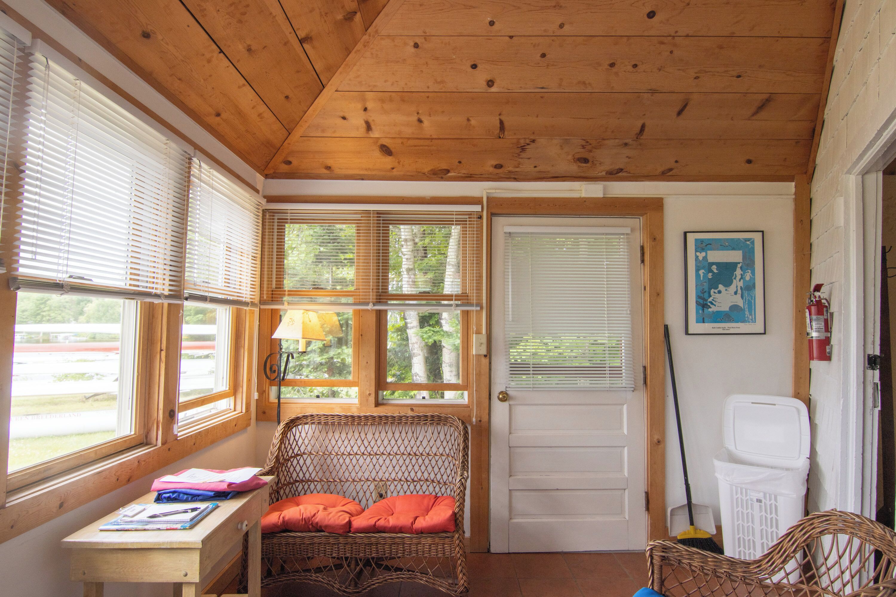 L’intérieur d’un chalet accueillant, avec un plafond en bois, un fauteuil en osier doté de coussins orange, un bureau avec une lampe et une porte avec une fenêtre.