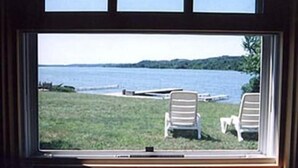 Blick aus dem Fenster auf eine Wiese mit zwei weißen Stühlen und einem Steg, der ins Wasser führt.
