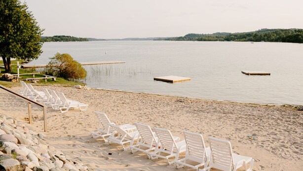 Ein Strand mit weißen Liegestühlen und einem hölzernen Steg, der ins ruhige Wasser führt.