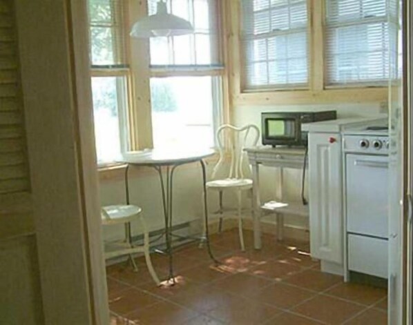 Una cocina con un refrigerador blanco, un microondas y una estufa. Hay una ventana con persianas y una silla cerca de la ventana.