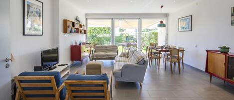 A spacious living area with a TV, sofa, chairs, a dining table, and a bookshelf.