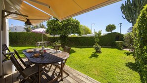 Eine Terrasse im Freien mit Tisch und Stühlen, einem gelben Sonnensegel und einem gepflegten Garten mit Hecken und Bäumen.