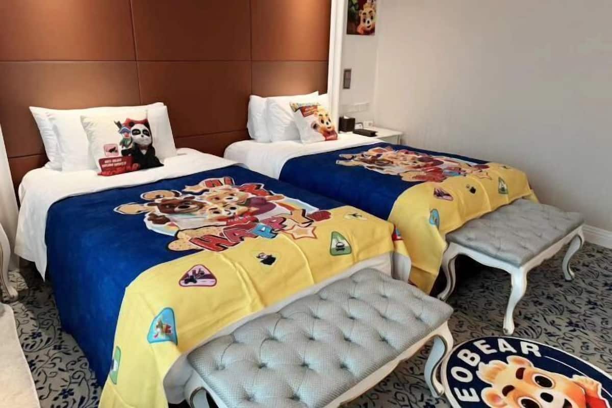 Ein Hotelzimmer mit zwei Betten, jedes mit einem cartoonartigen Bettbezug, einer grau gepolsterten Bank und einem runden Teppich mit Bärenmuster.