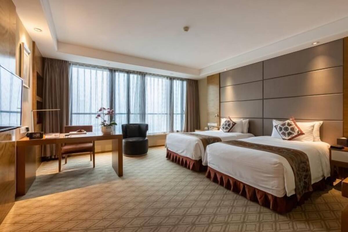 Ein Hotelzimmer mit zwei Betten, einem Schreibtisch, einem Stuhl und einem großen Fenster mit Vorhängen.