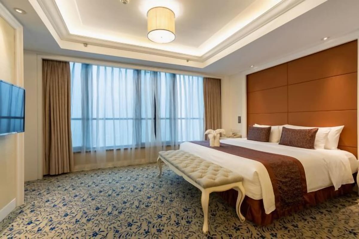 A hotel room with a large bed, a bench, a flat-screen TV, and a large window with curtains.