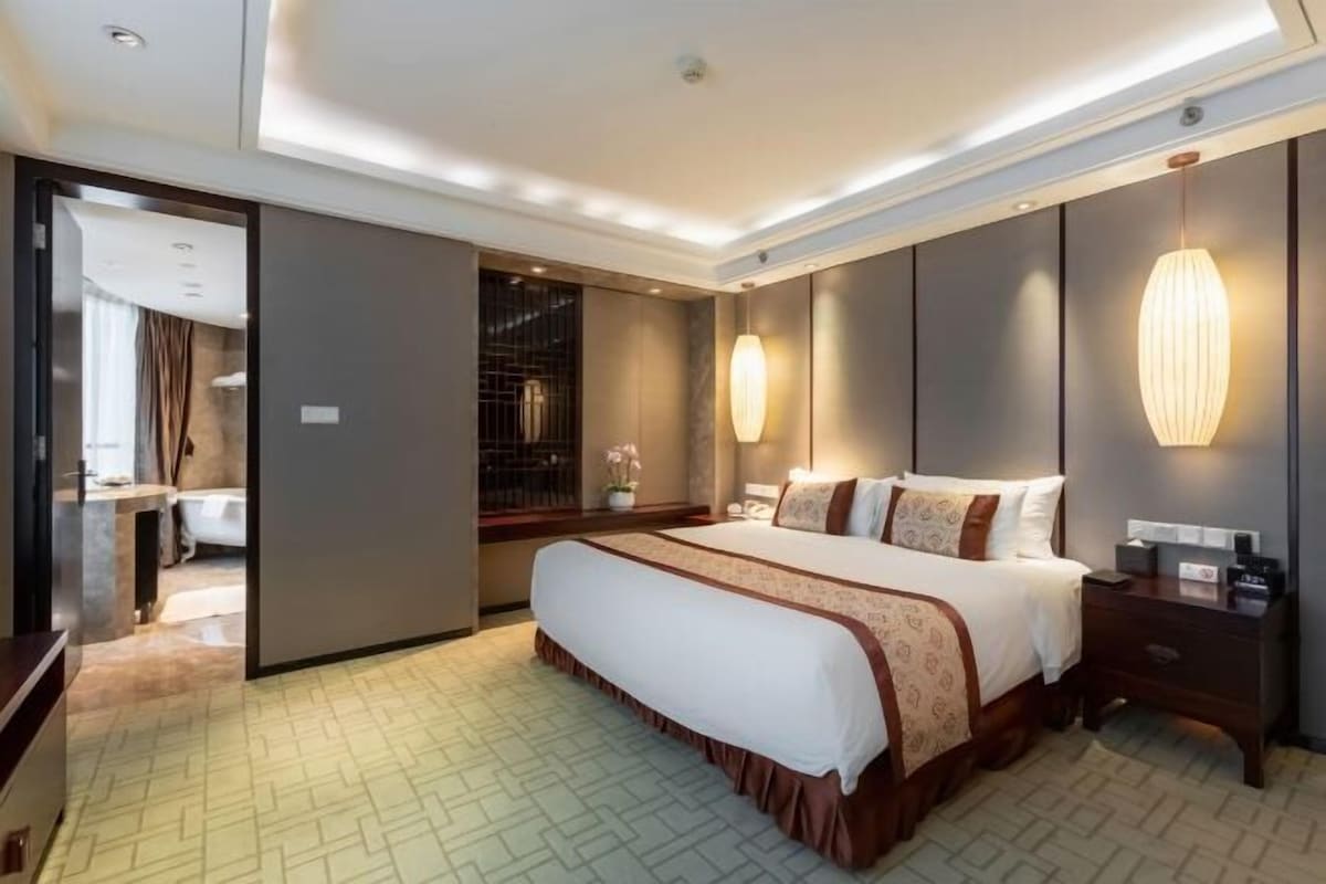 A hotel room with a large bed, bedside tables, a nightstand, a mirror, and a wall-mounted lamp.