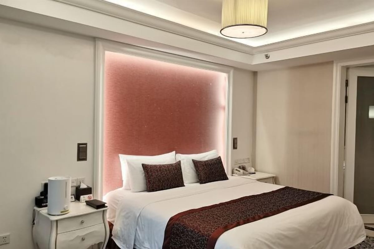 A hotel room with a large bed, a bedside table with a lamp, and a door with a glass panel.
