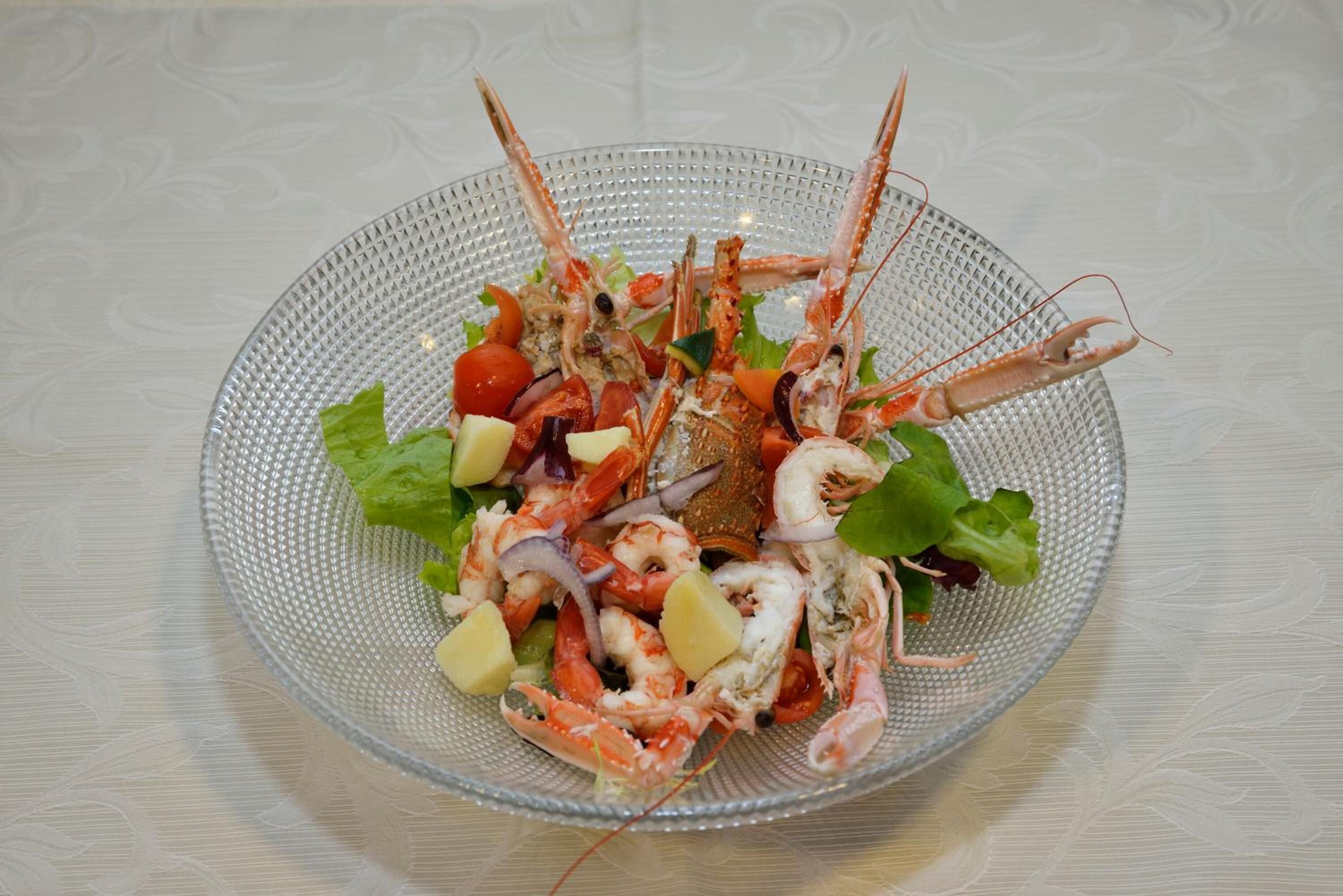 A glass bowl containing a seafood salad with various vegetables and a whole lobster.