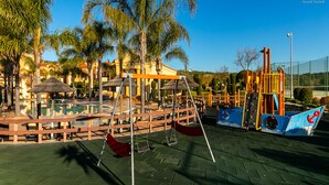 A playground with swings, a climbing structure, and a pool area surrounded by palm trees and a fence. - A24 - LuzBay Beach Apartment by Dreamalgarve (Lagos)