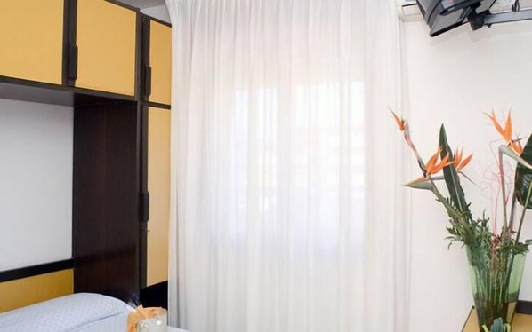 Une chambre d’hôtel avec un lit, une fenêtre ornée de rideaux légers et une plante en pot avec des fleurs de strelitzia.