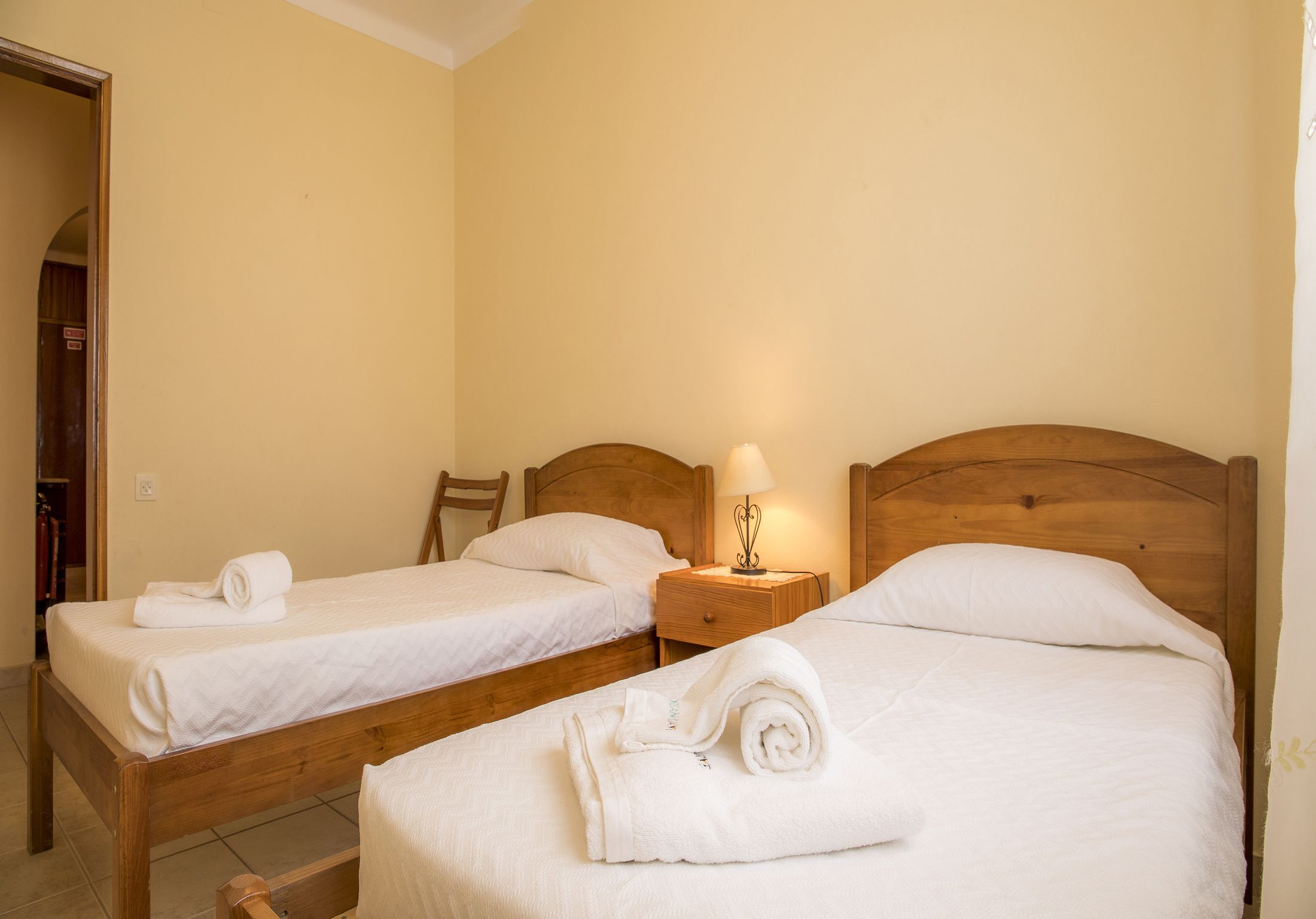 Two single beds with wooden headboards, a nightstand with a lamp, and a rolled towel on each bed.