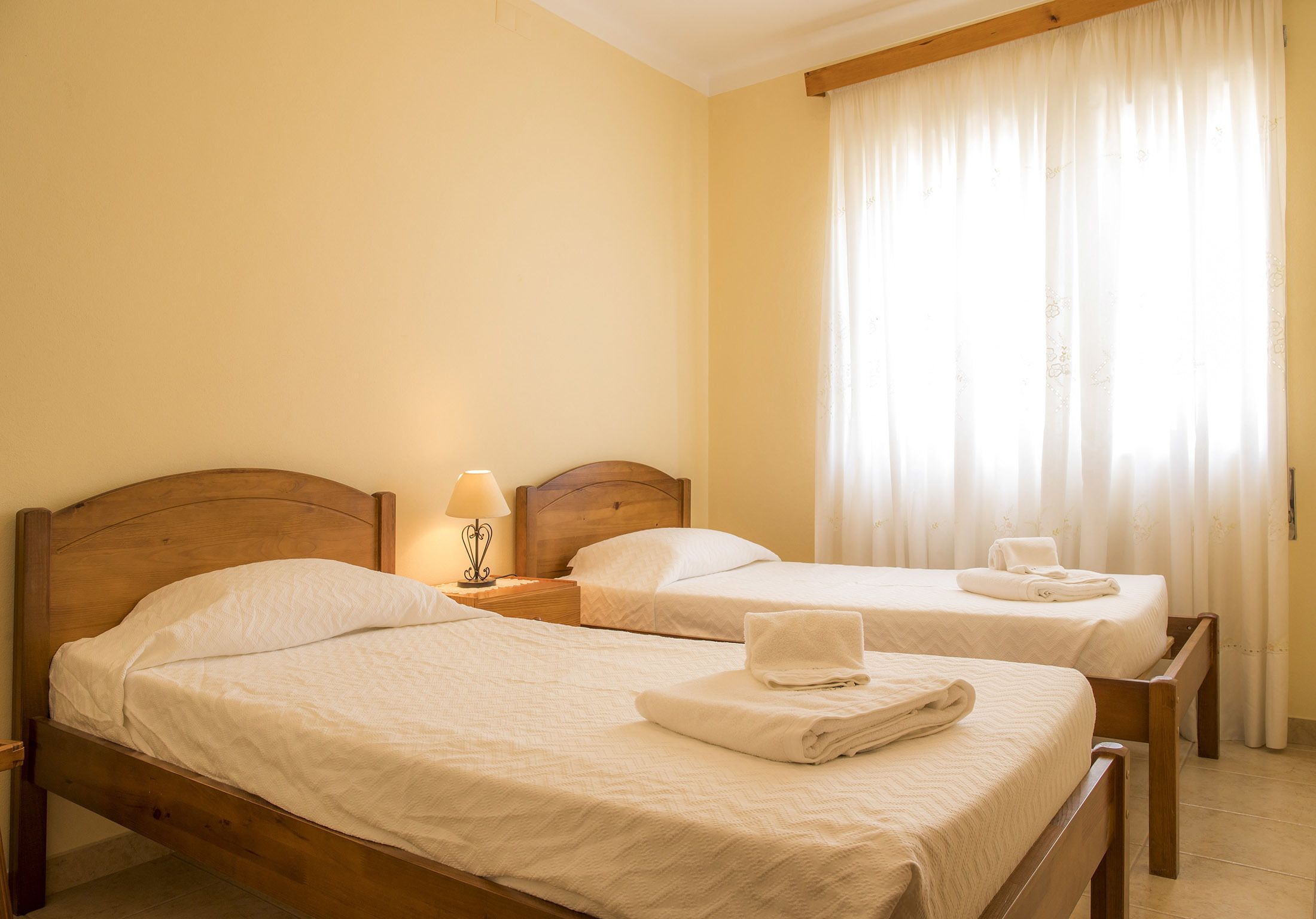 Dos camas individuales con estructuras de madera y ropa de cama blanca, una mesita de noche con lámpara y una ventana con cortinas de gasa.